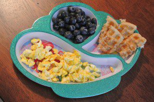 A meal in a divided plate including scrambled eggs, a waffle and blueberries. 