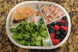 A meal in a divided plate including broccoli, a rice cake with hummus, chicken breast and raspberries and blueberries. 