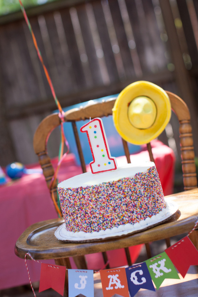 A photograph of a cake with a number one candle set on the tray of a highchair.