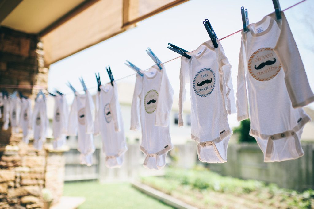 A row of onesies hanging from a clothesline. Each onesie has a patch labeled with a consecutive number of months, ending with 11 months. 