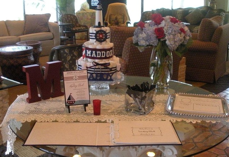 A table on top of which is a cake with a nautical theme, a vase of flowers, a block of the letter M, markers in a glass container, a framed invitation and a binder. 