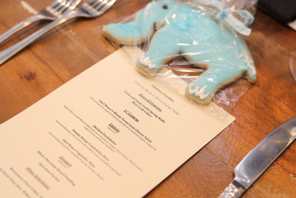 A menu listing Hors d'oeuvres, first course, entrée, desert and wine selections. A blue elephant cookie wrapped in cellophane with a ribbon is placed at the top of the menu. 