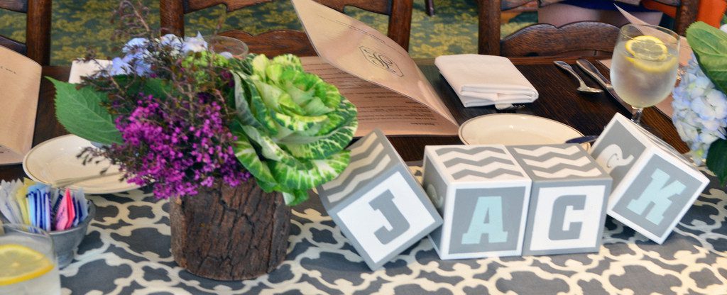 A centerpiece on a table with a flower arrangement in a wooden vase and blocks arranged to spell Jack all on top of a table runner. 