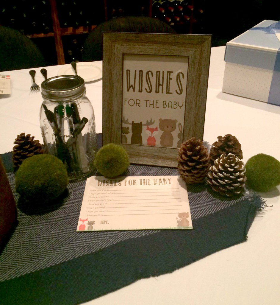 A table centerpiece consisting of a framed text stating wishes for the baby, a card titled wishes for the baby, pinecones, a jar of markers, and green fluffy balls all on top of a table runner. 