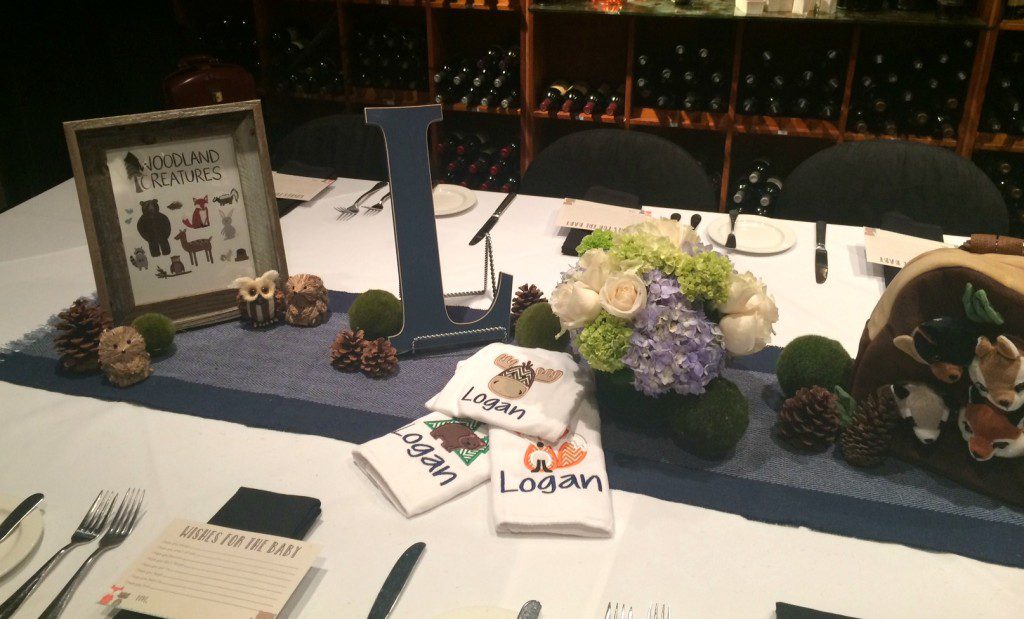 A centerpiece on a table with a flower arrangement, A large block L, small ornamental animals, pinecones, and three cloths each with embroidered animals and the name Logan all on top of a table runner. 