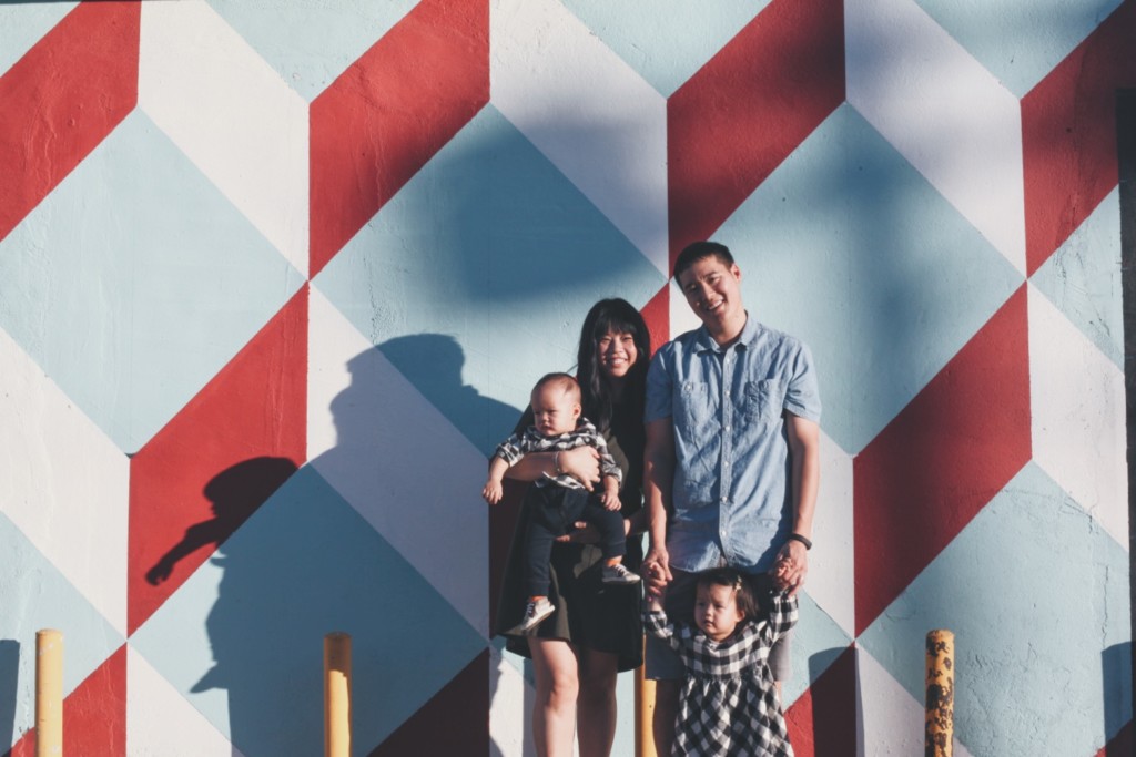 A father holding the hands of a toddler and a mother holding a baby while standing in front of a mural. 