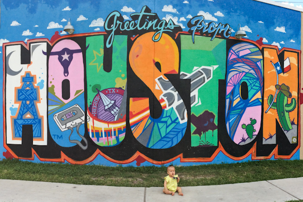 A baby sitting in front of a mural with the text Greetings from Houston. 