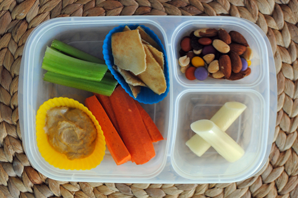 A plastic container with three divisions containing a trail mix, a cheese stick, celery sticks, carrot sticks, pita bread and hummus. The pita bread and hummus are in muffin cups. 