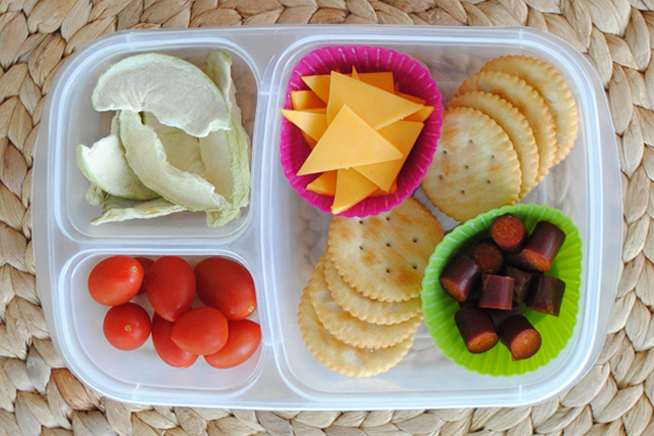 A plastic container with three divisions containing freeze dried apples, grape tomatoes, crackers and cheese and sausage. The cheese and sausage are in individual muffin cups. 