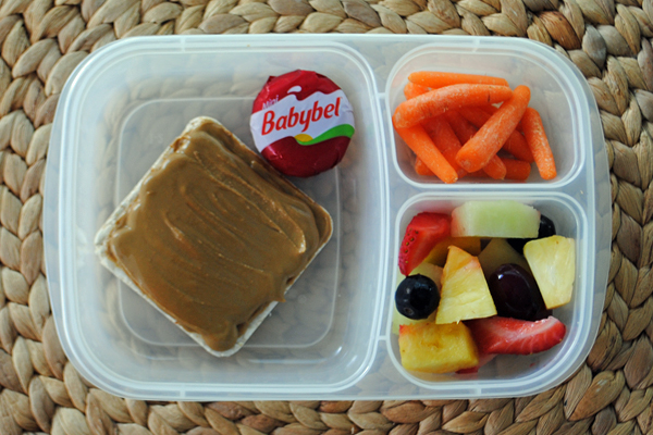 A plastic container with three divisions containing a rice cake with sunbutter, a piece of babybel cheese, a fruit salad and carrots.