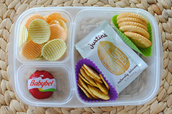 A plastic container with three divisions containing veggie Chips, a babybel cheese, a package of peanut butter, crackers and banana chips. The banana chips and crackers are in muffin cups. 