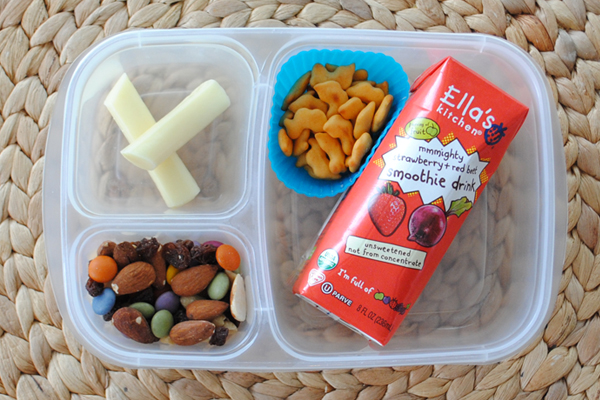A plastic container with three divisions containing trail mix, a cheese stick, a smoothie drink and a muffin cup filled with Quack N Bites. 