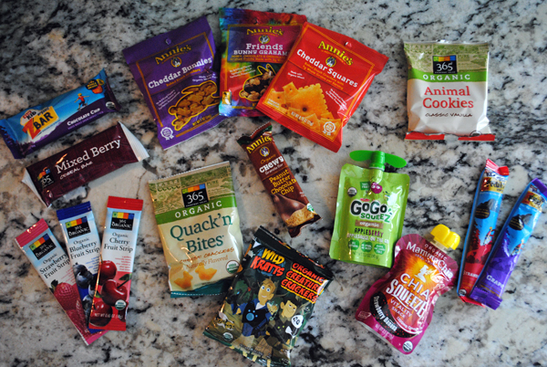A photograph of 16 snacks including A z bar, a mixed berry bar, three types of annie's snacks, three types of fruit strips, Quack N Bites, a trail bar, animal cookies, a chia squeeze, organic creature crackers and two fruit packages. 