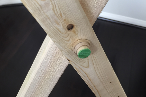 A wooden dowel extends through a hole at the point where the two wood boards cross to form a V. 