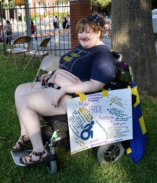 A teenage girl with a nasal canula and in a wheelchair with a sign on the side that states: Make PRIDE accessible to everyone. 