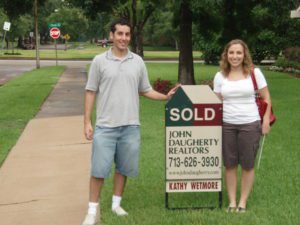 A photograph of a husband and wife standing next to a Sold real estate sign. 