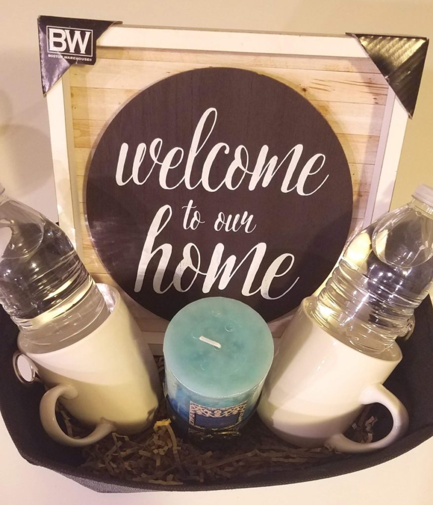 A welcome basket containing a candle, two mugs, two bottles of water and a sign reading welcome to our home. 