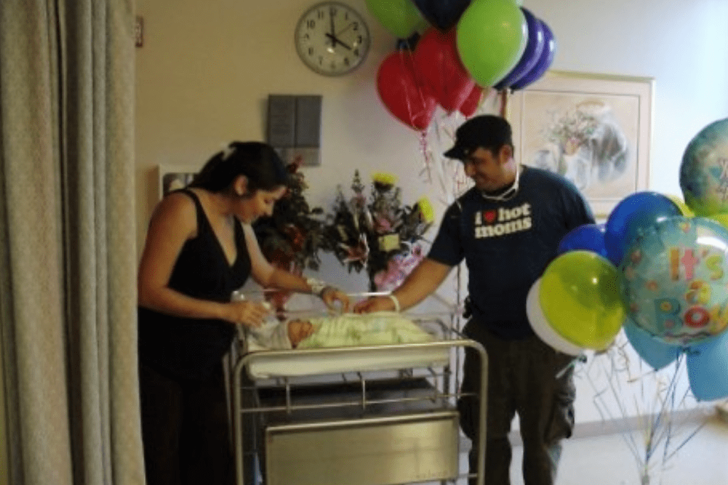 A photograph of a man and a woman smiling and standing over a newborn baby at a hospital. Balloons with the text It's a boy are nearby. 