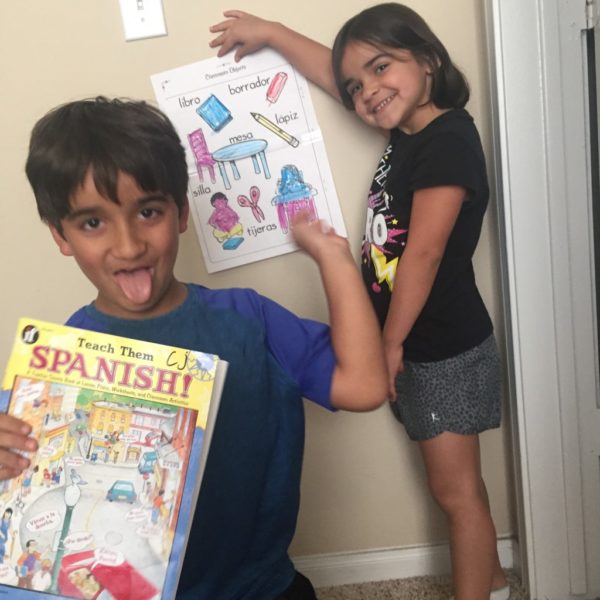 Two young children. The boy holds a book titled Teach them Spanish. The girl holds up a page of Spanish vocabulary words.