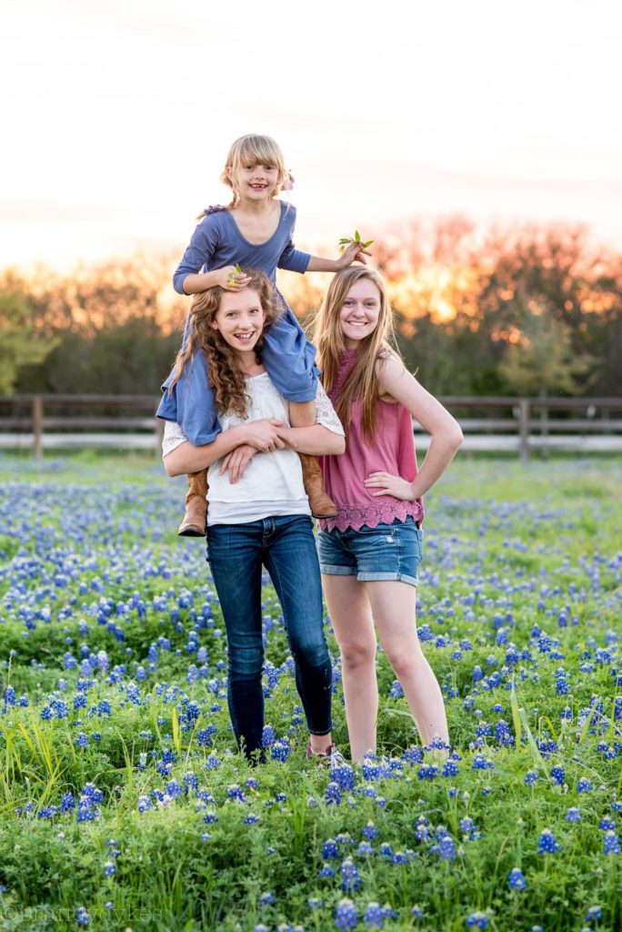 Two smiling teenagers standing in a field of bluebonnets.  One teenager holds a younger girl on her shoulders. 