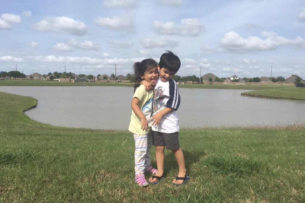 Two smiling children with their arms around each other standing next to a pond. 