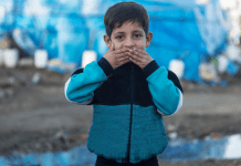 A refugee child pressing his hands to his lips.