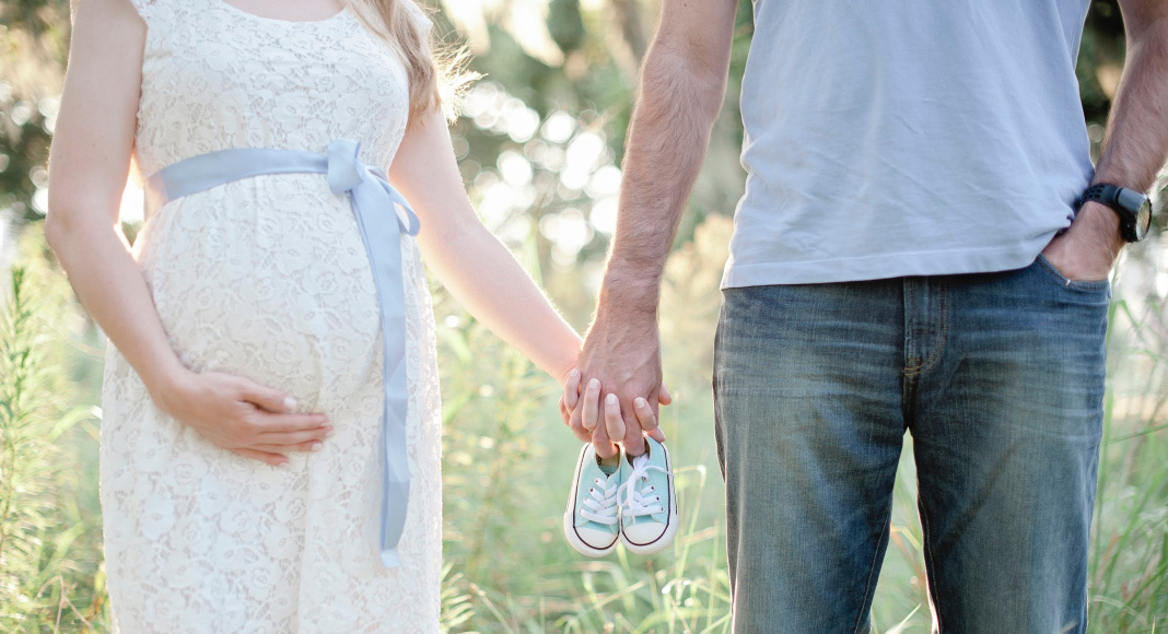 A pregnant woman and a man holding hands and holding a pair of baby shoes.