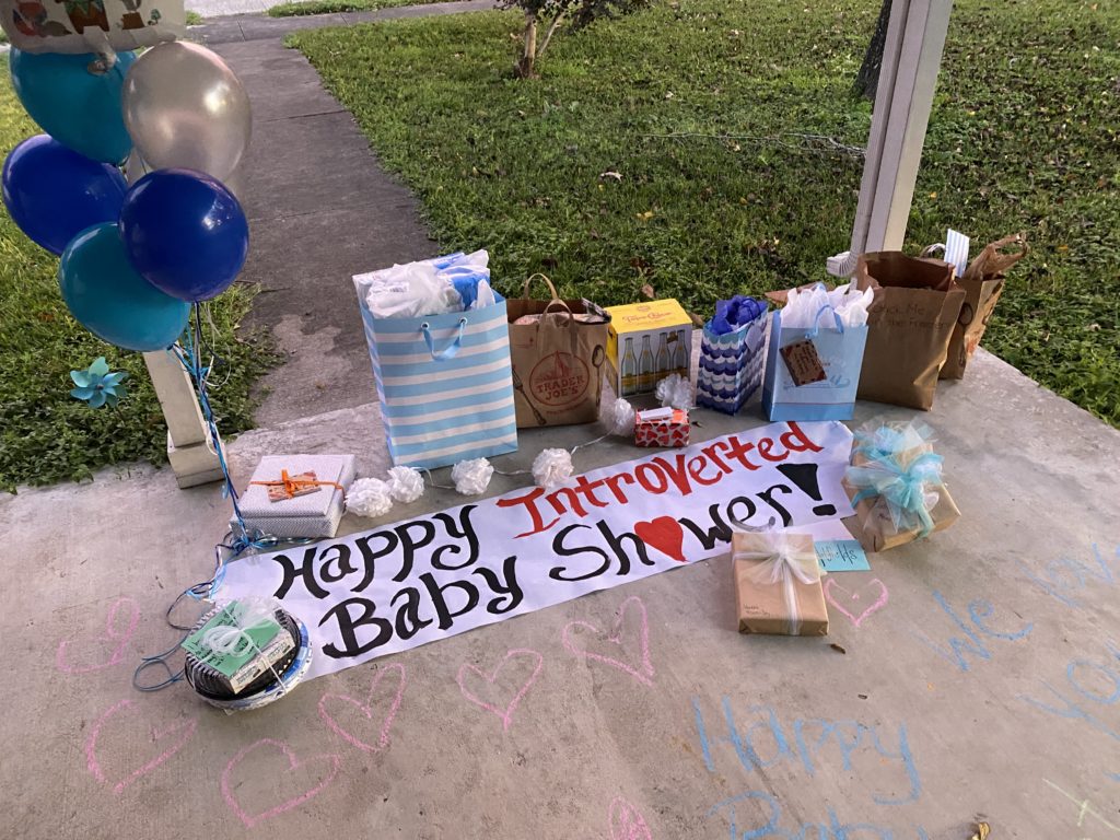 Wrapped gifts, helium balloons and a sign with the text Happy Introverted Baby Shower, all on the ground on a porch. 