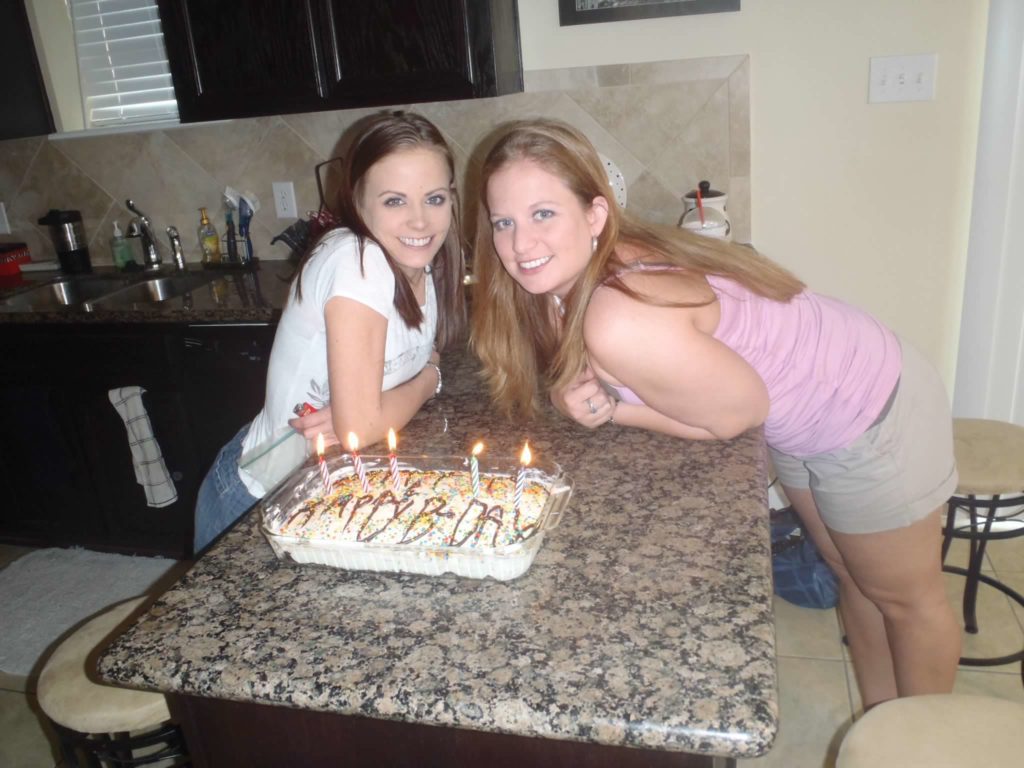 A photograph of two smiling women standing at a counter behind a birthday cake with lit candles. 