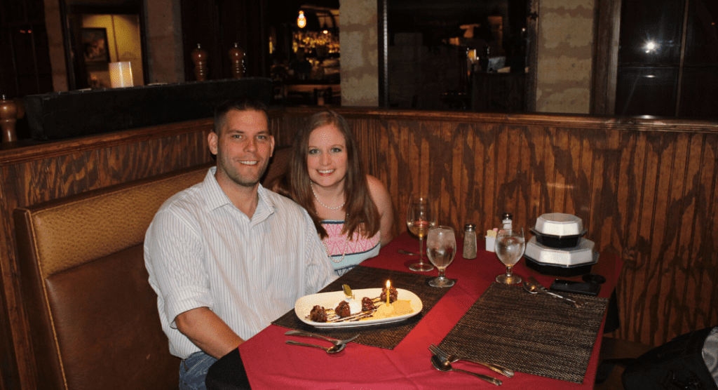 A photograph of a husband and wife at a restaurant with a dessert with a candle in front of him. 