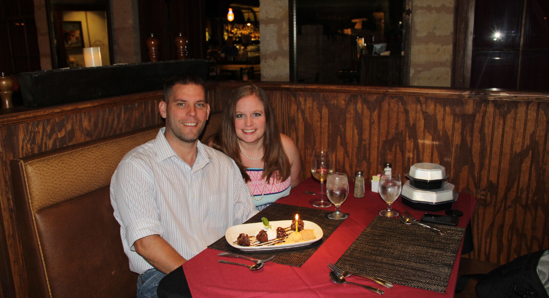 A photograph of a husband and wife sitting at a restaurant with a dessert with a lit candle in front of him.