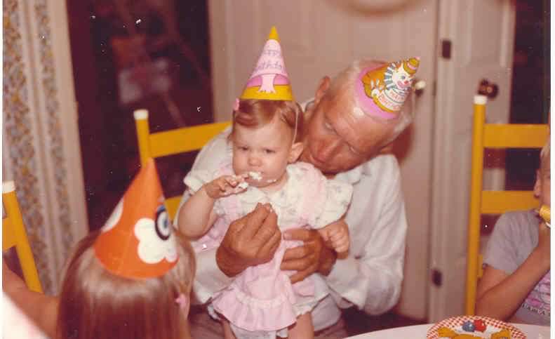A photograph of a toddler standing on her father's lap and eating cake at a birthday party. 