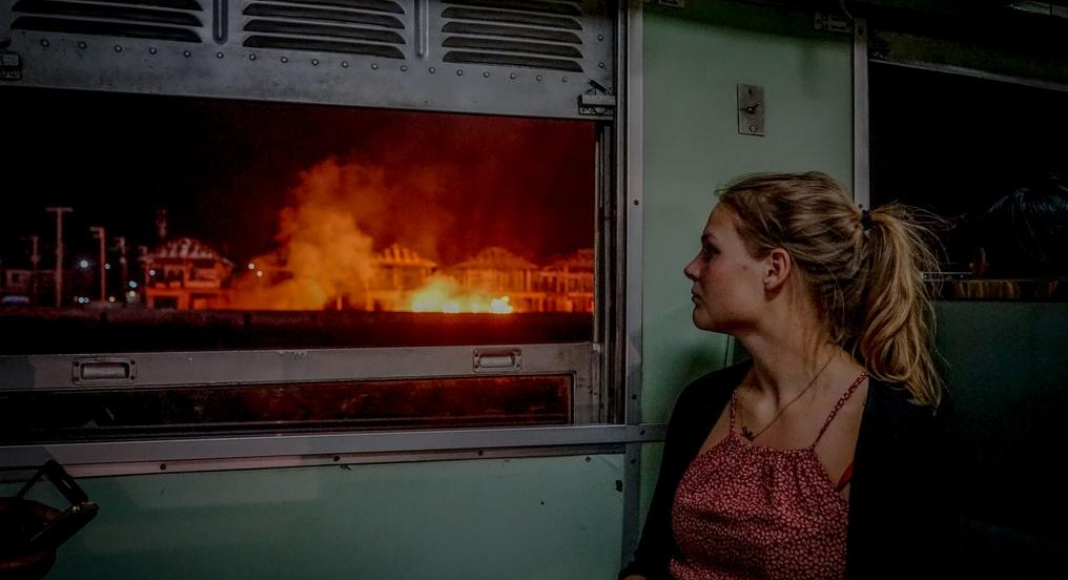 A photograph of a woman looking out a window at a house on fire.