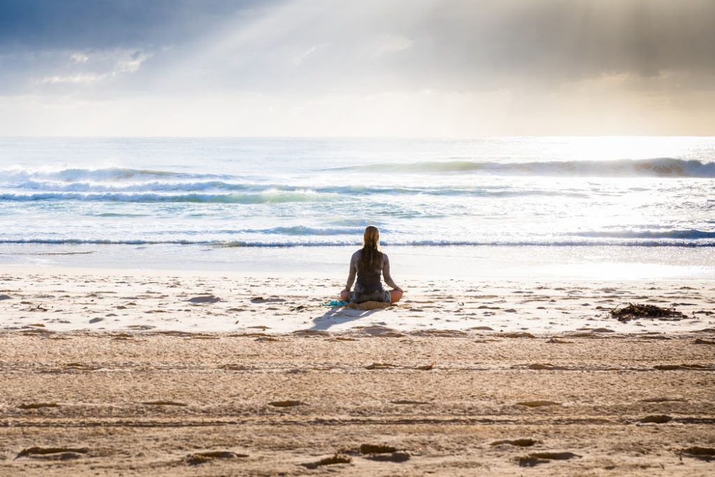 A woman sitting cross-legged on a beach and facing the ocean. 