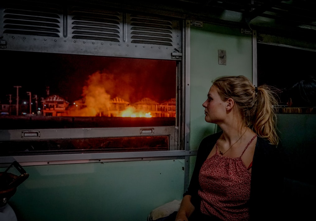 A woman staring out a window at a house on fire. 