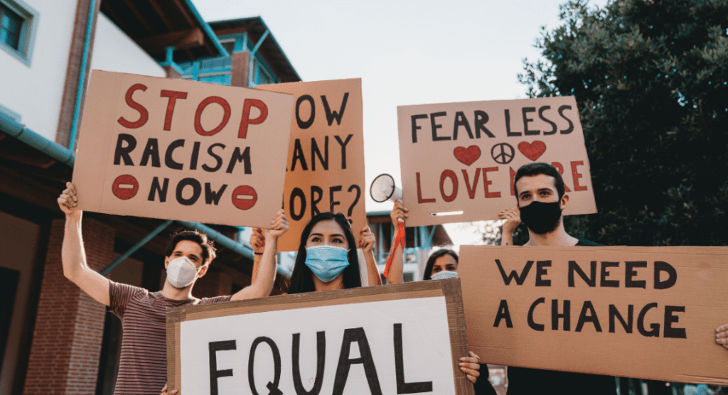 A group of People holding signs with the text: Stop Racism Now. Fear Less Love more. We need a change. 