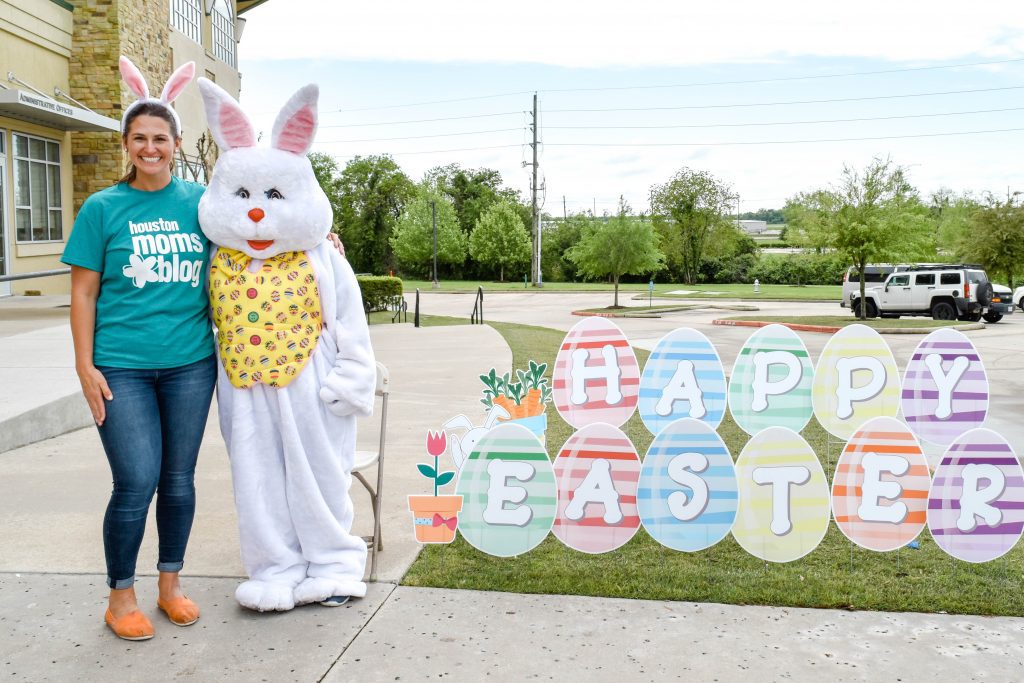 A woman wearing a Houston Moms blog T-shirt and bunny ears posing with the Easter bunny next to a Happy Easter sign. 