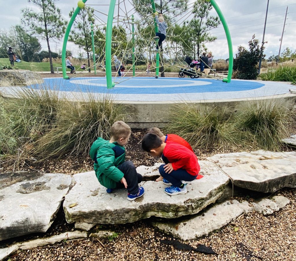 Three young children exploring a hole in a rock with a playground in the background. 