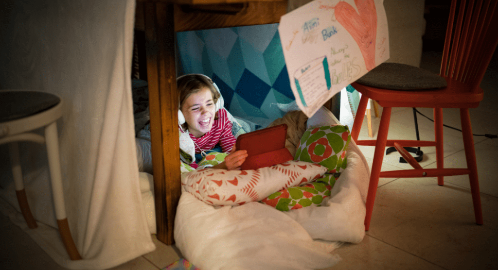 A child with headphones on in a tent made of blankets in a house.