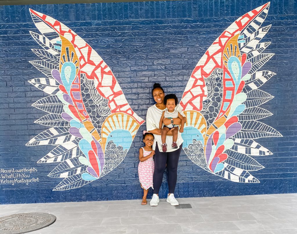 A mother holding a child and standing next to another child while standing in front of a mural with wings. 