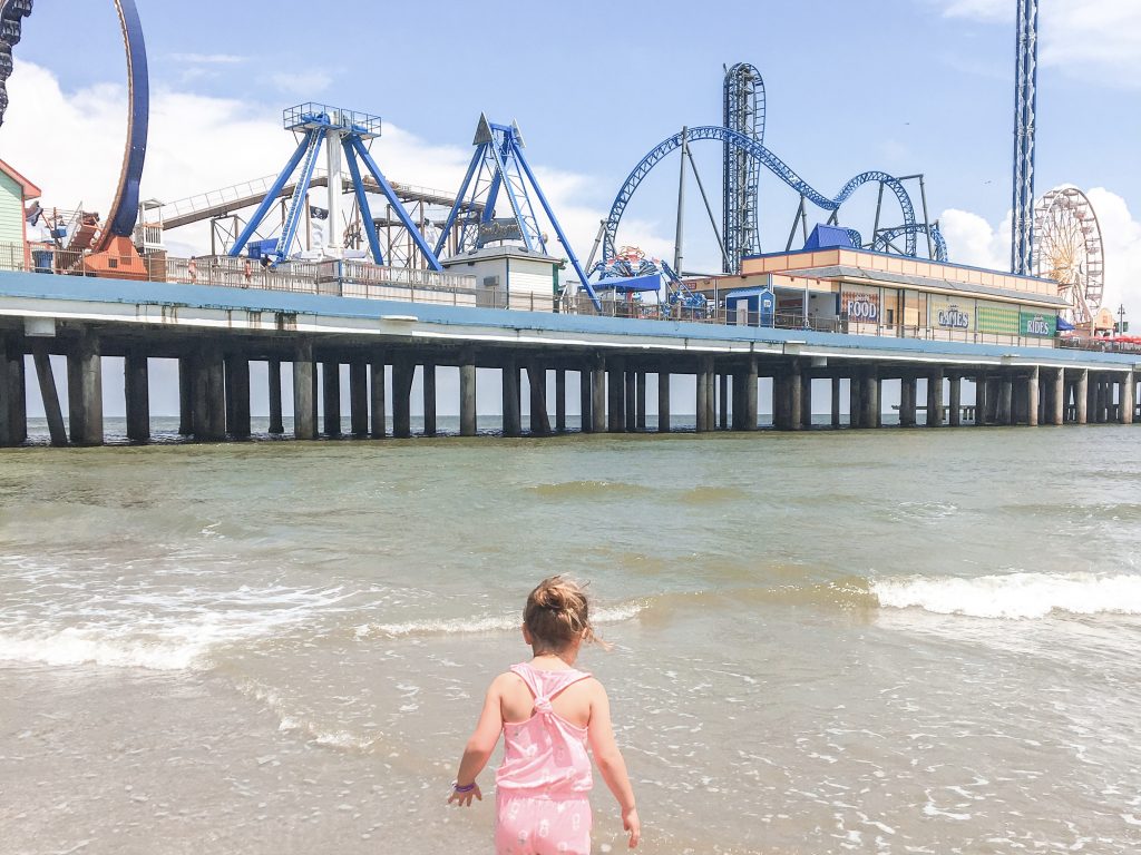 A child stands in the water at a beach with an amusement park in the background. 