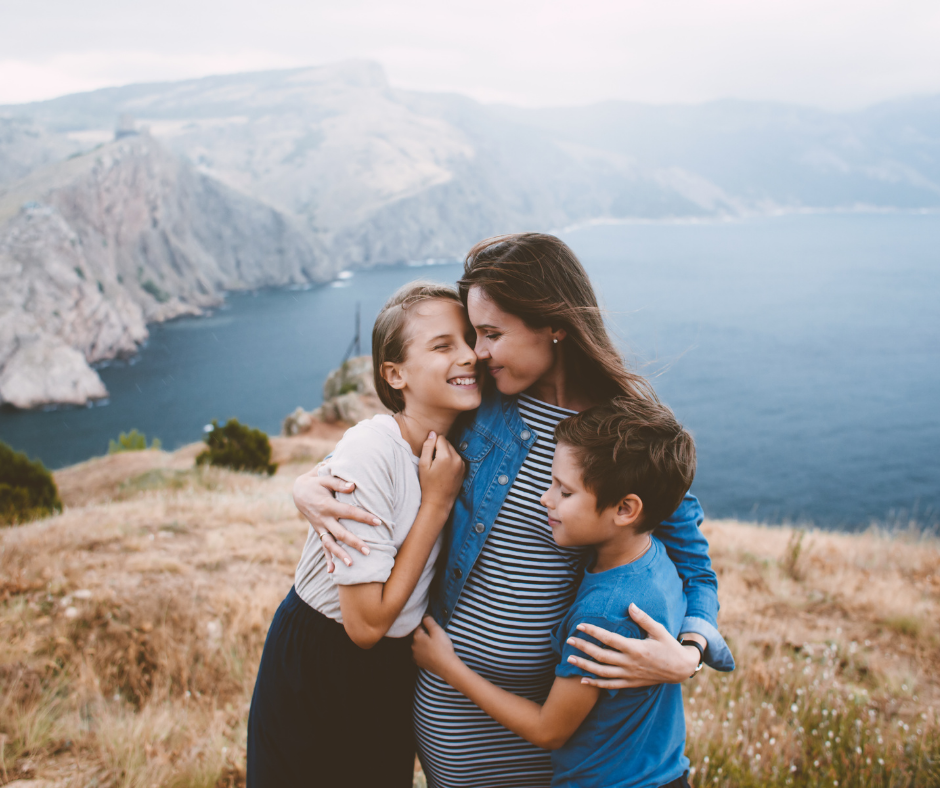A mother hugging her two children on a hill overlooking a body of water. 