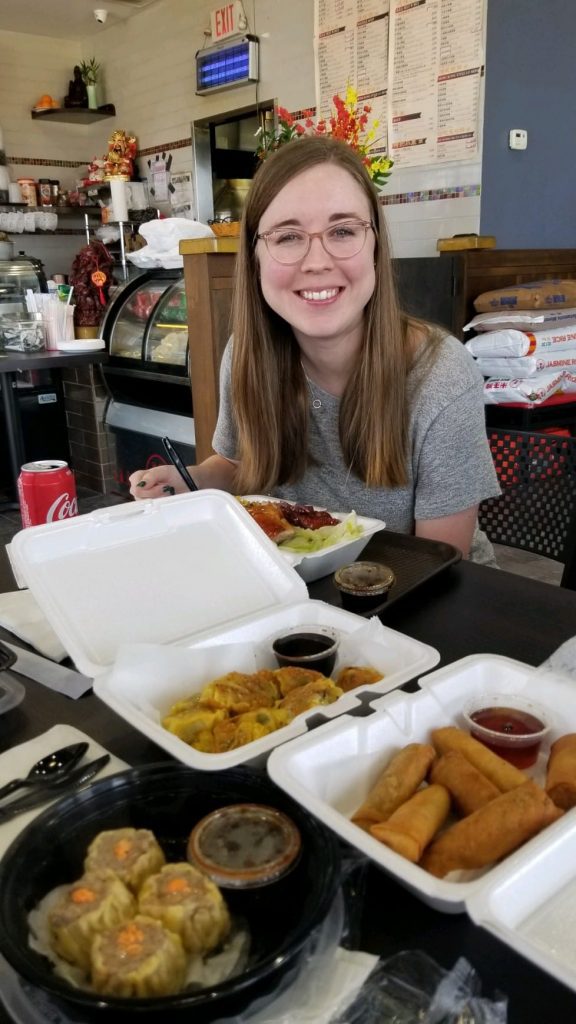 A smiling woman sitting at a restaurant with Asian food on the table. 