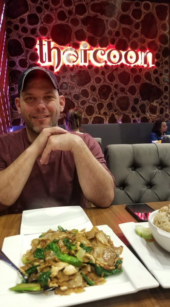 A smiling man sitting at a restaurant table with a dish of Asian food in front of him. A sign in the background reads Thaicoon. 