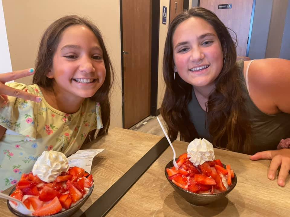 A child and a teenager sitting at a restaurant table with two bowls of shaved ice topped with strawberries and whipped cream. 
