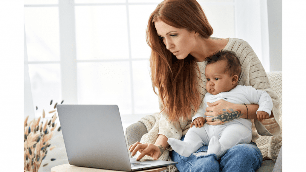 A mother holding a baby and typing on a laptop. 