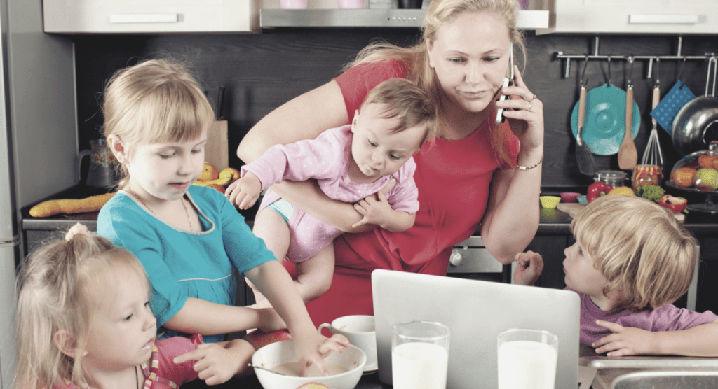A photograph of a mother holding a baby with three other children next to her at a table. She is on the phone and looking at a laptop. 