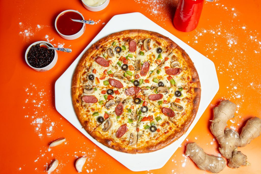 a supreme pizza on white tray with organge background surrounded by sauces
