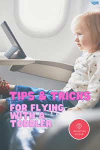 Tips and Tricks for Flying with a Toddler. A photograph of a toddler sitting on an airplane seat watching an ipad on the tray table. Logo: Houston moms. 