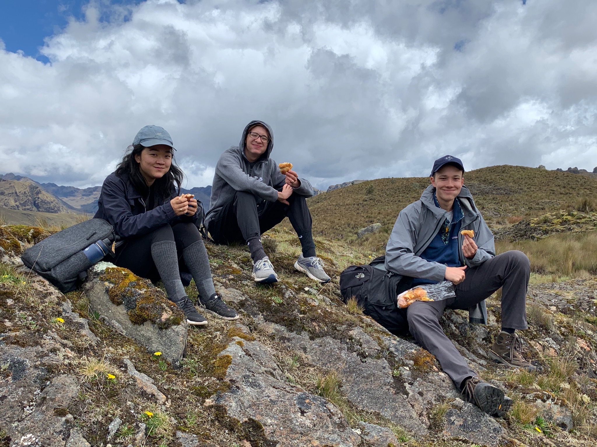 3 teens on AMIGOS trip sit on a mountain eating a snack